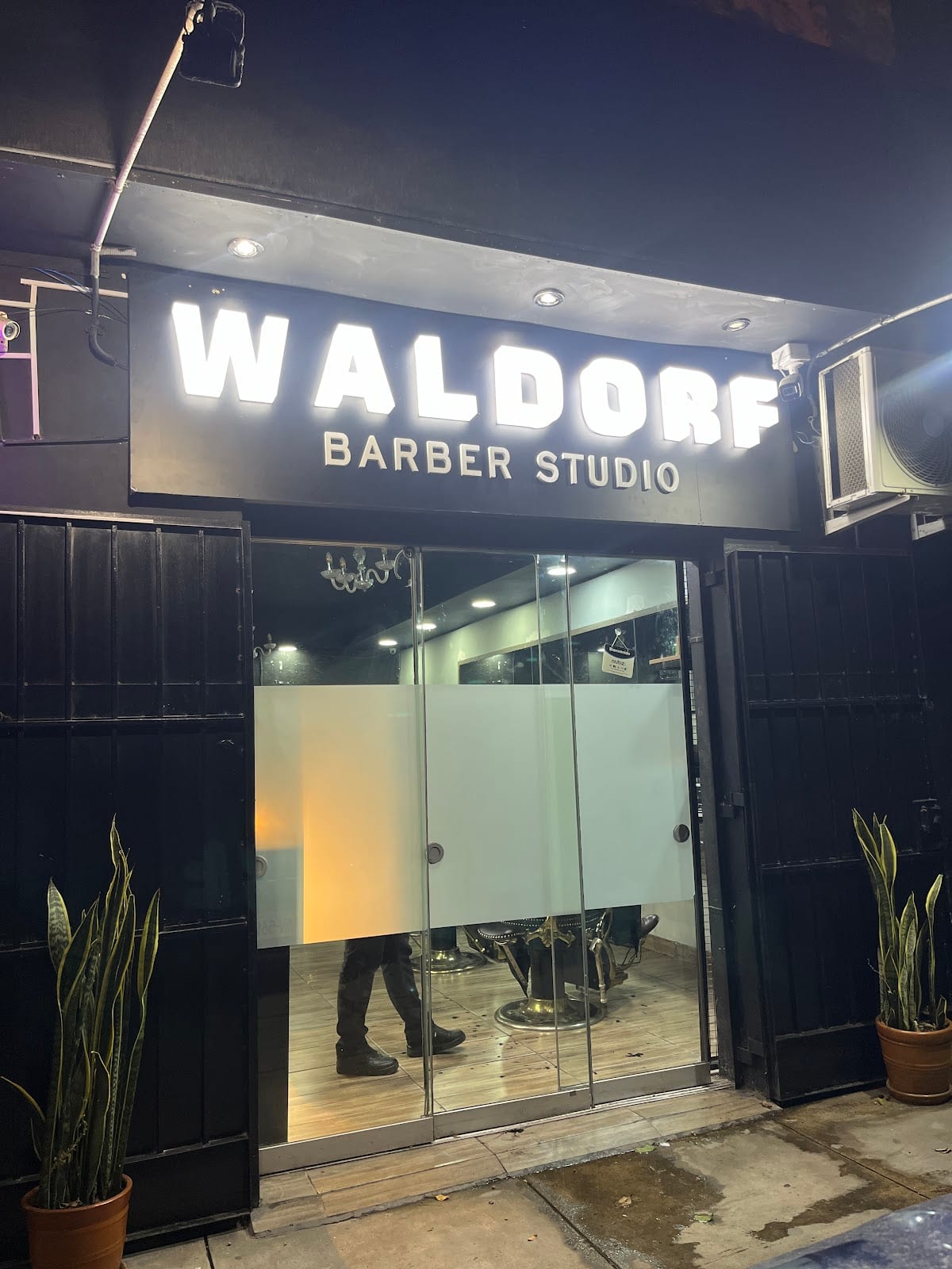 Fachada de Waldorf Barber Studio iluminada de noche en Av. Militar